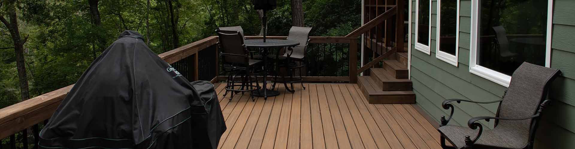 Wooden deck with grill, table, chairs, and house with windows. Forest background.