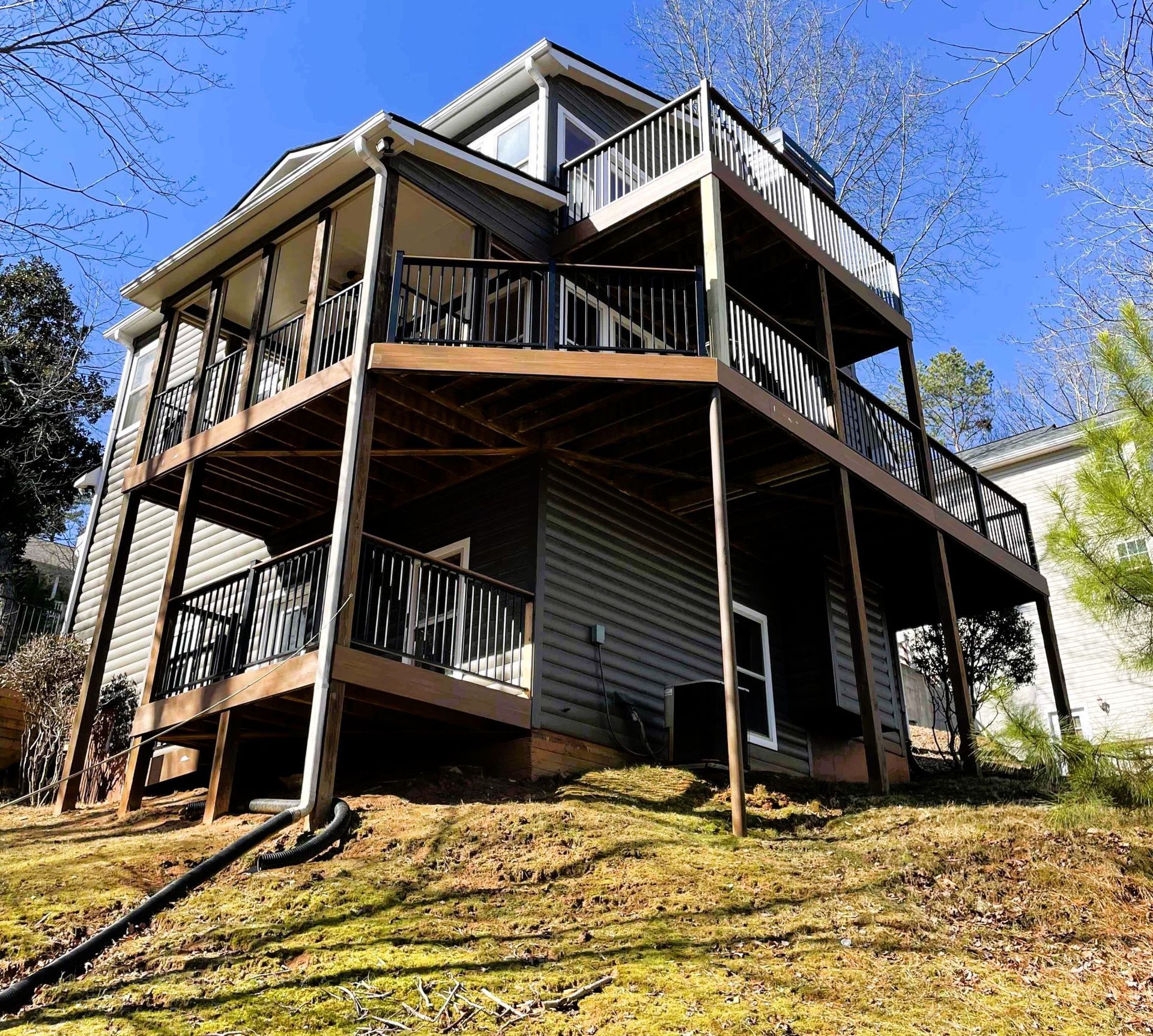 Two-story house with tiered wooden decks, gray siding, and black railings, on a grassy hillside.