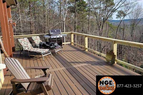 Wooden deck with grill, chairs, and view of trees.