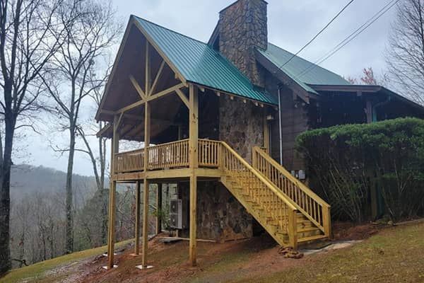 Cabin with wooden deck and staircase. Green metal roof, stone chimney, surrounded by trees.