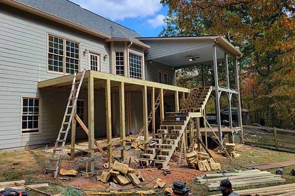 Deck construction in progress at a beige house.  Wooden framing and stairs are visible with lumber and tools nearby.