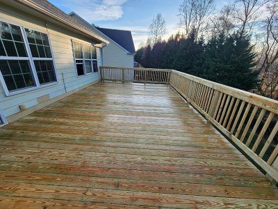 Wooden deck attached to a house with a railing, trees, and sky in the background.
