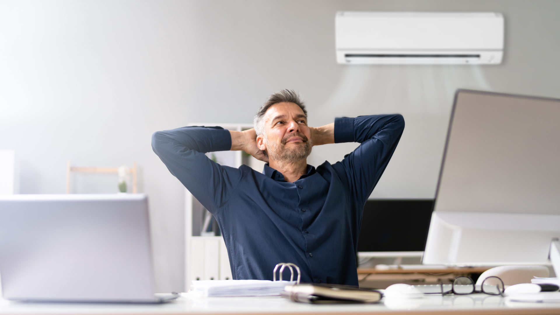 A man is sitting at a desk with his hands behind his head under an air conditioner.