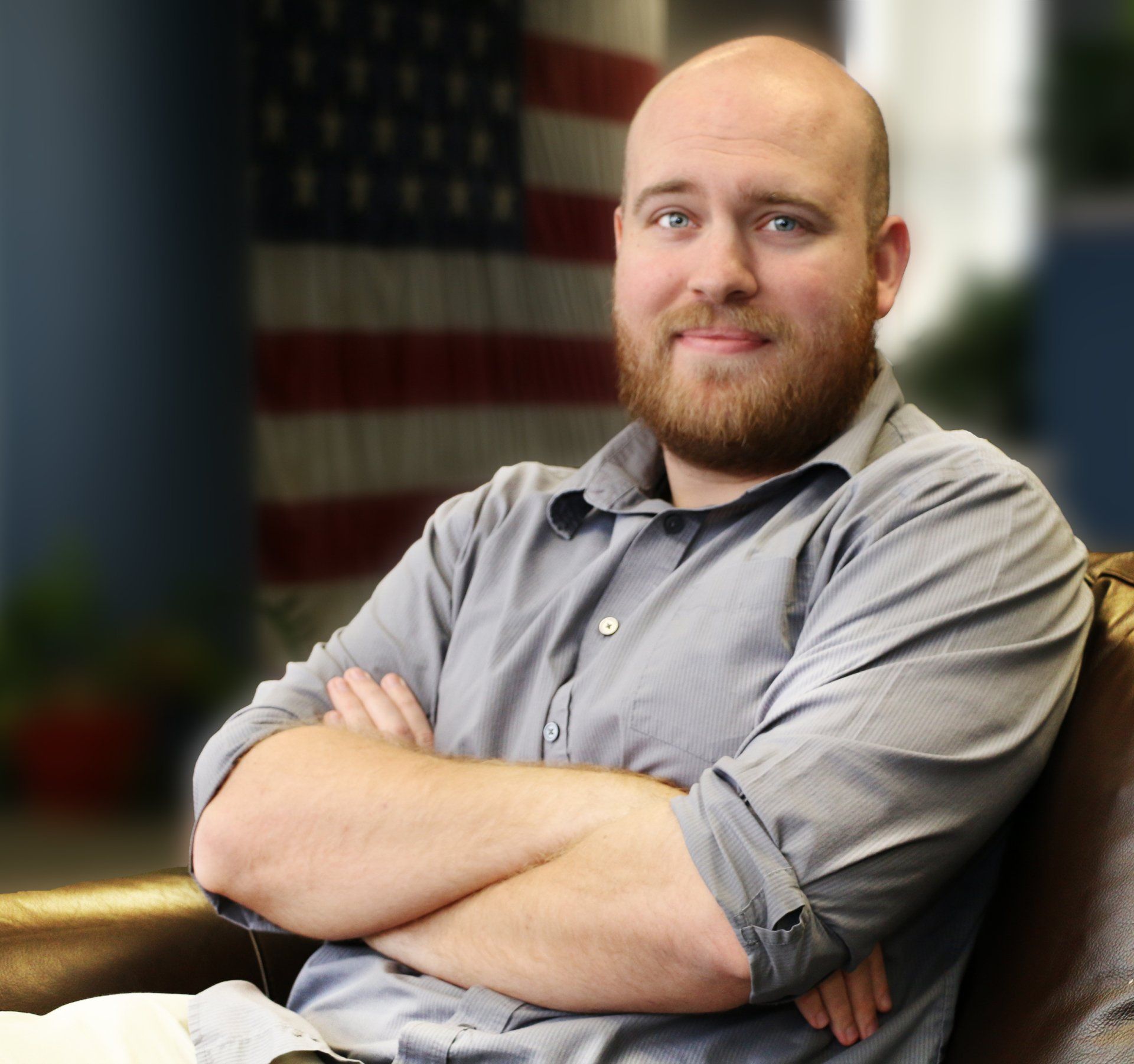 A bald man with a beard is sitting on a couch with his arms crossed