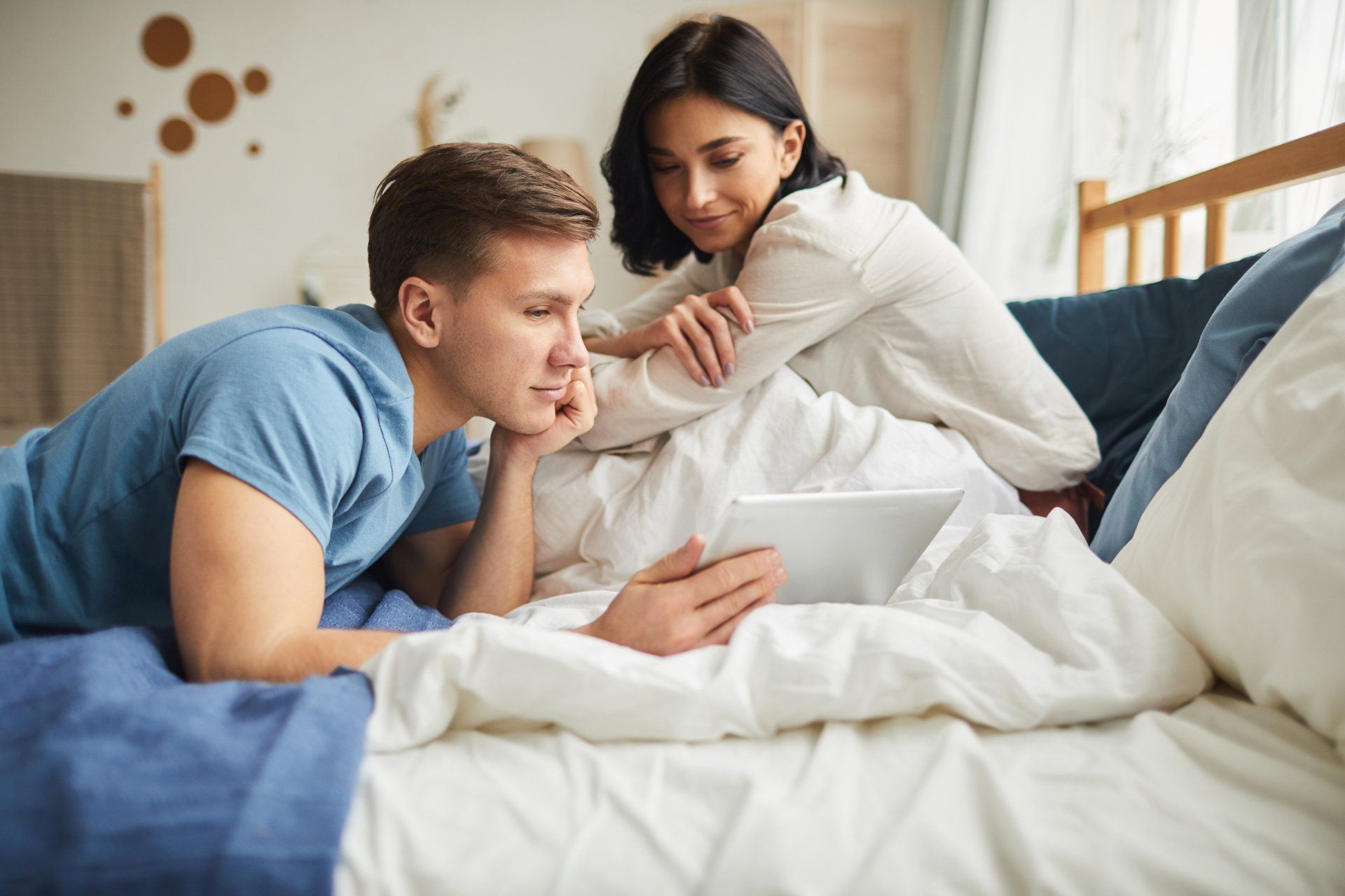 A man and a woman are laying on a bed looking at a tablet.