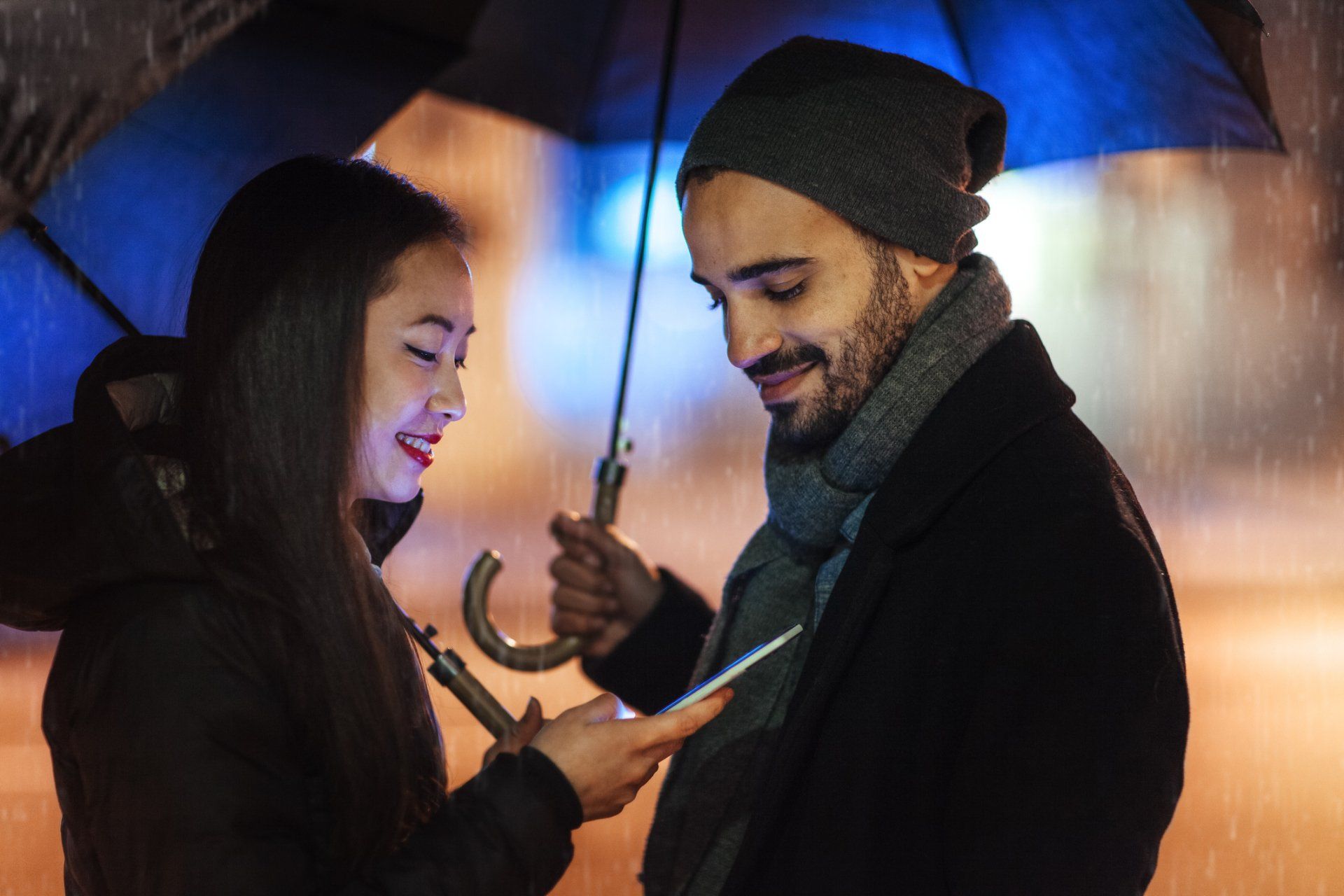 A man and a woman are standing under an umbrella looking at a cell phone.