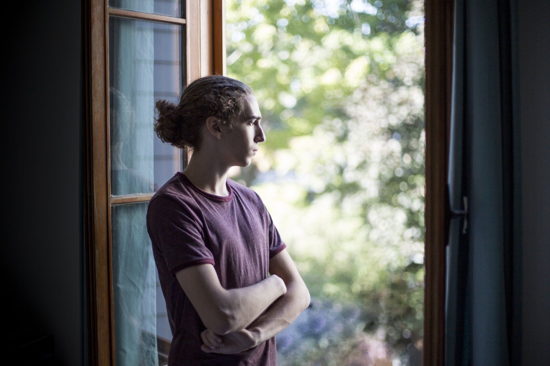 A young man is standing in front of a window looking out.