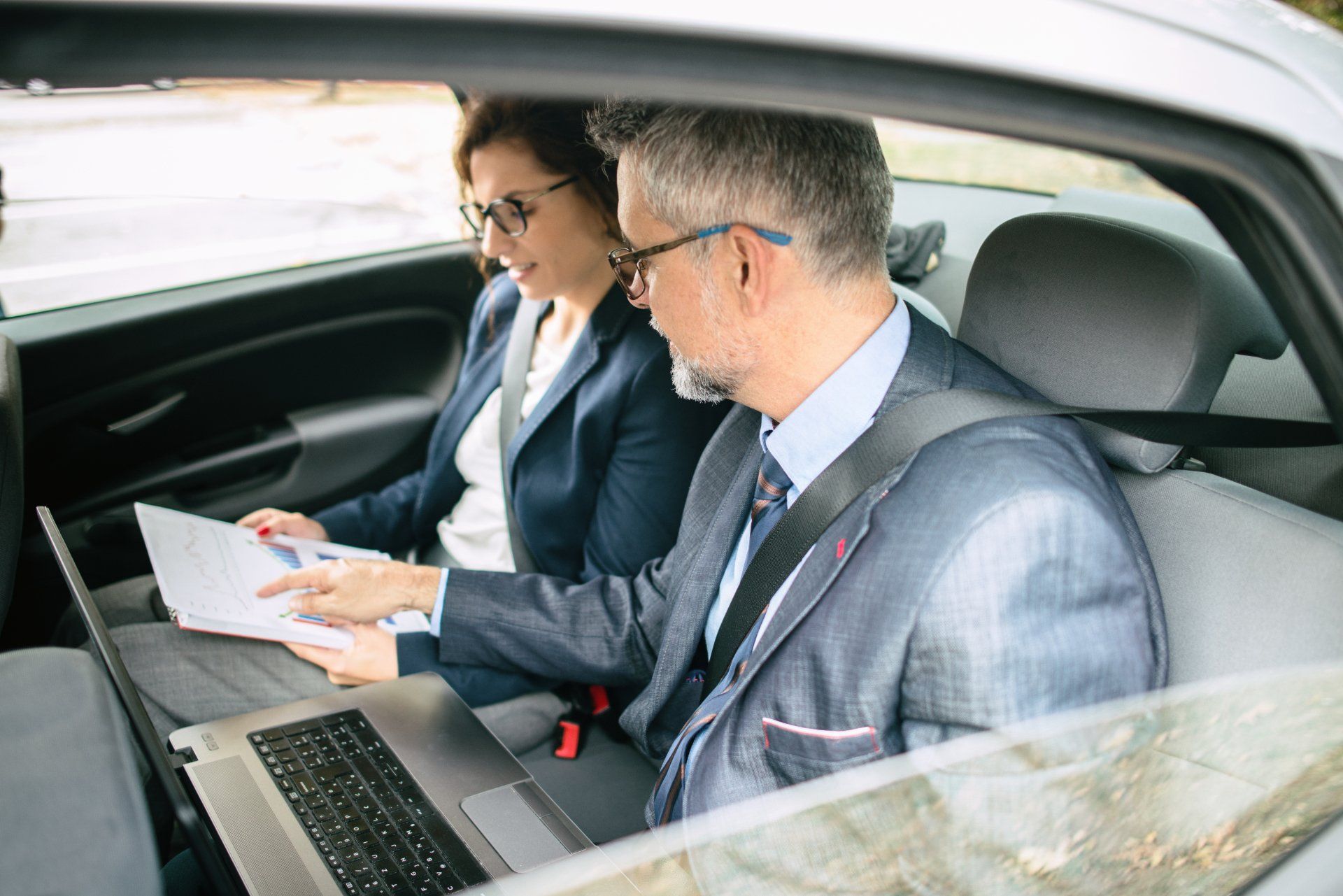 A man and a woman are sitting in the back seat of a car looking at a laptop.