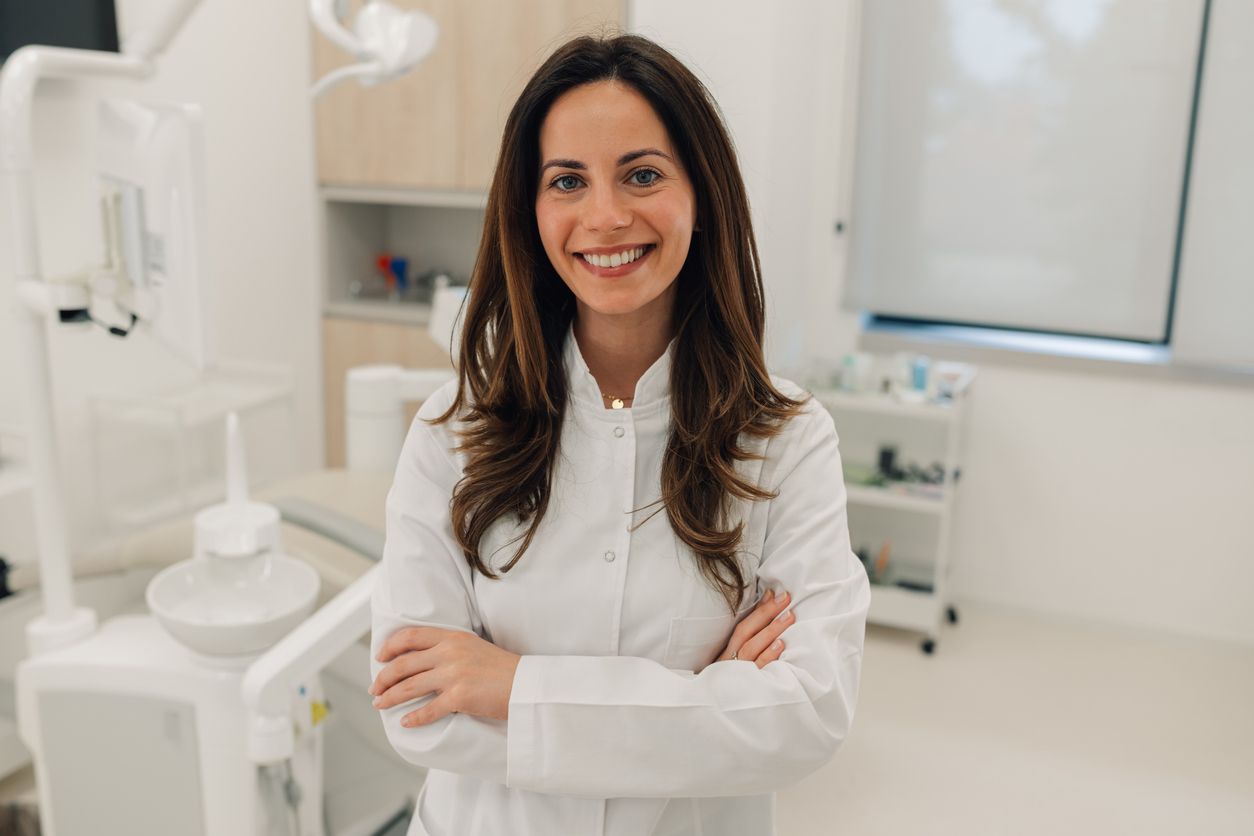 Woman in lab coat smiles, arms crossed, in a dental office.