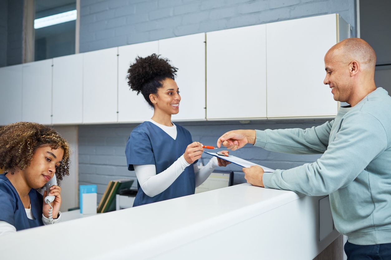 Medical staff assisting a patient at a reception desk. One worker hands paperwork to the man.