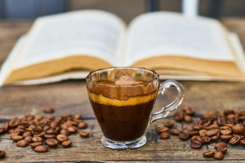 Espresso in glass cup with coffee beans on wood table, open book in the background.