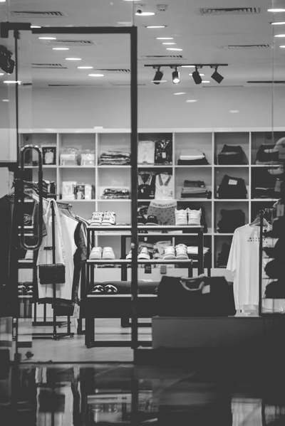 Zwart-witfoto van het interieur van een kledingwinkel met schappen, rekken en hangende kledingstukken.