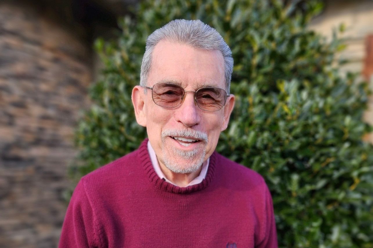 A man wearing glasses and a red sweater is standing in front of a bush.