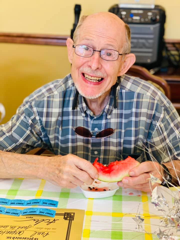 An elderly man is sitting at a table eating a slice of watermelon.