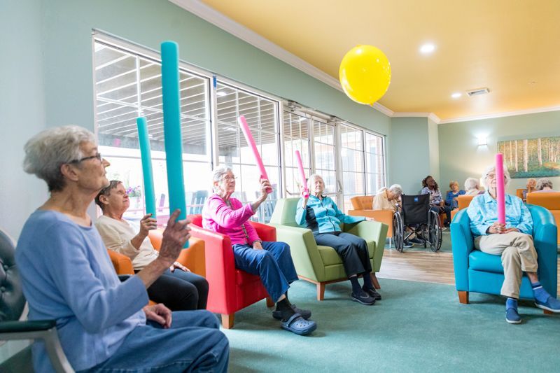A group of elderly people are sitting in chairs holding foam sticks in a living room.
