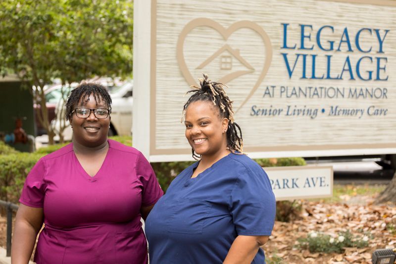 Two nurses are standing in front of a sign for legacy village at plantation manor.