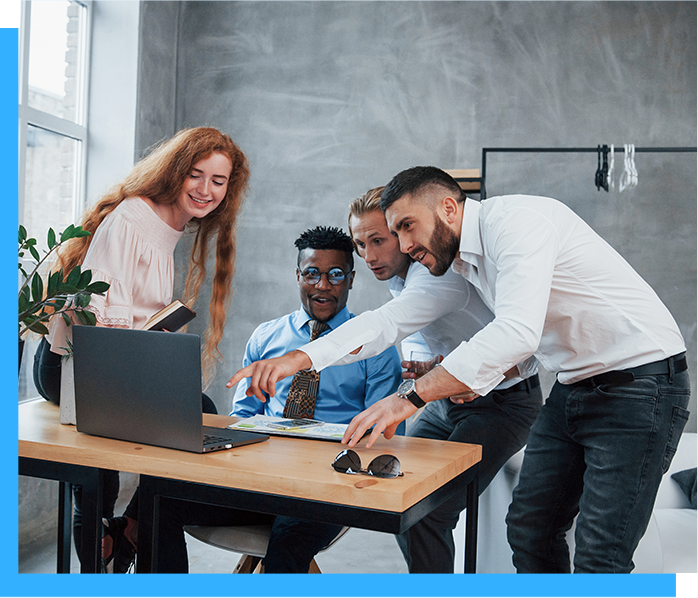 A diverse group of office workers gather around a laptop, pointing and discussing. They are in a modern office with a window and minimalist decor.