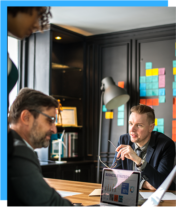 Business team in a meeting, reviewing a presentation on a laptop. Dark-paneled office, colorful sticky notes on the wall.