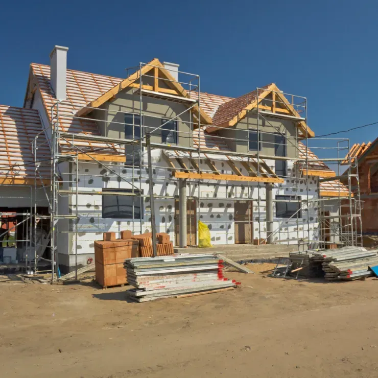 House under construction with scaffolding, insulation, and roof tiles. Exterior view under a blue sky.