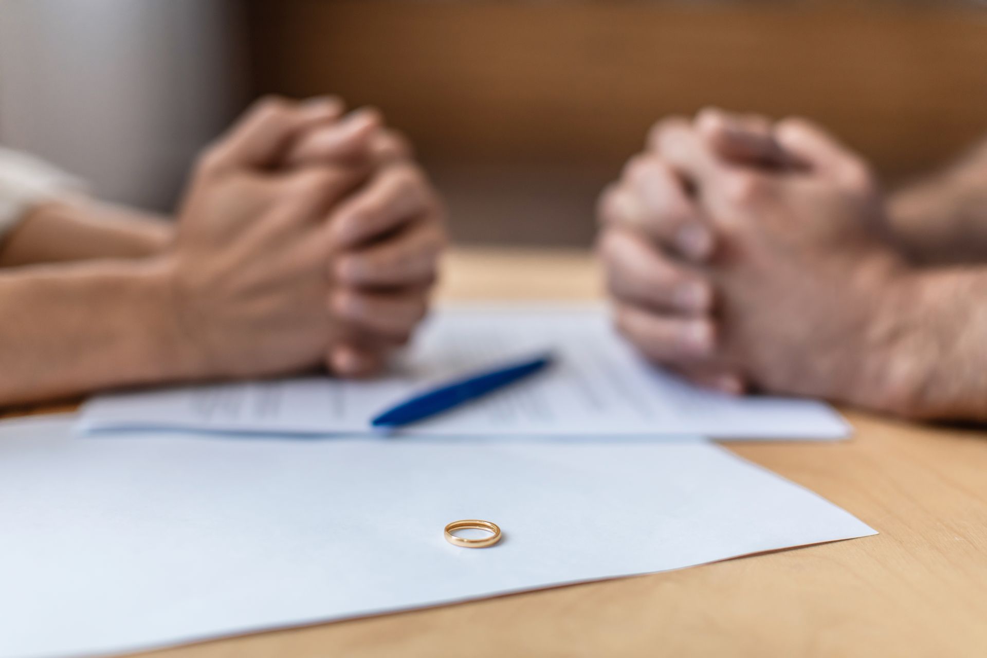 Hands clasped on a table, divorce papers, wedding ring; blurred couple in background.
