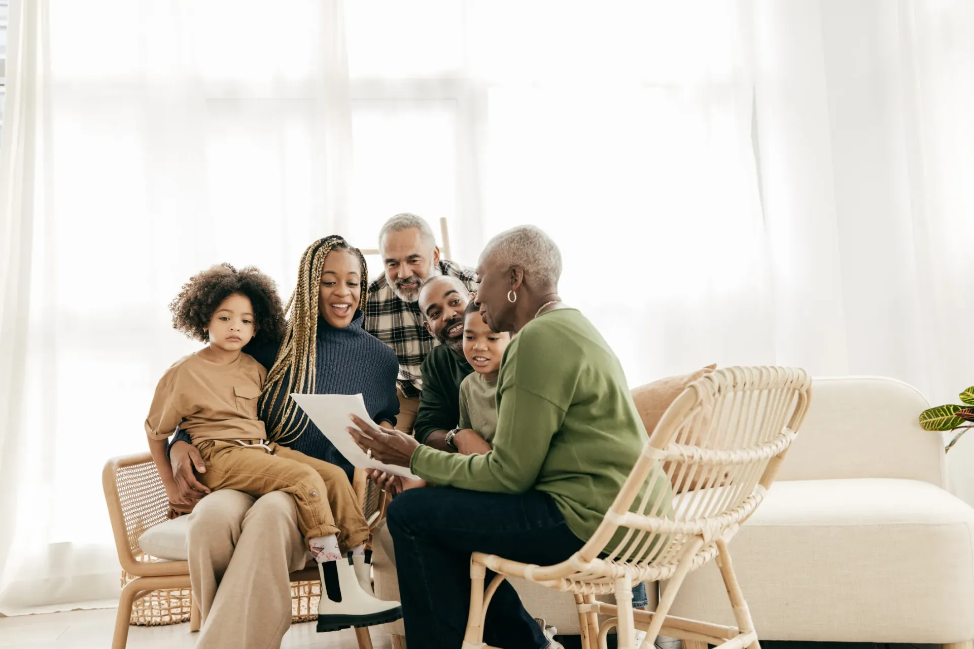 A multi-generational Black family sits together, looking at a document. The setting is a bright living room with a wicker chair.