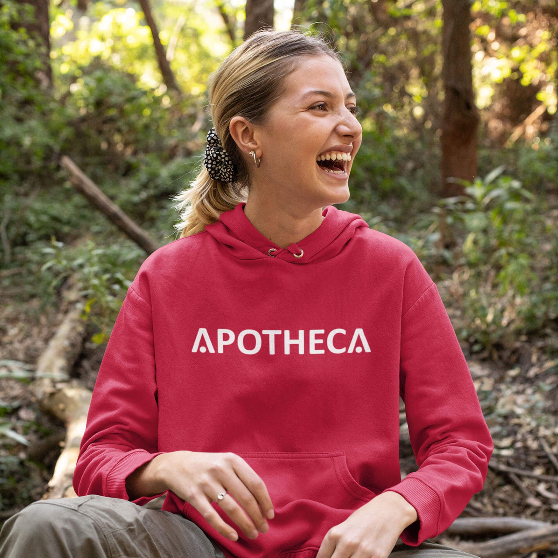 A woman wearing a red hoodie with the word apotheca on it is sitting in the woods.