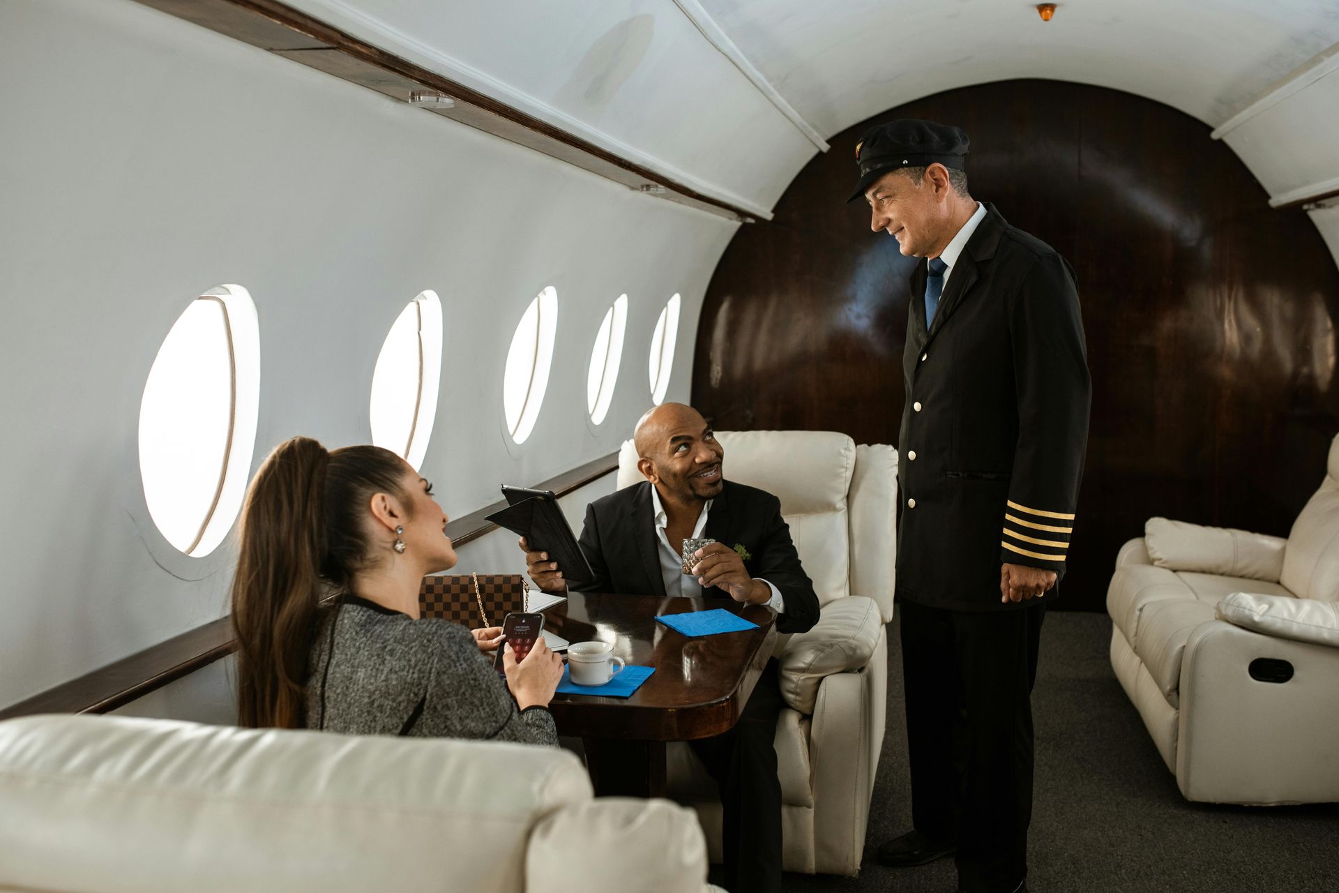 Pilot talking to two passengers in a private jet cabin. The passengers, a man and woman, are seated at a table.
