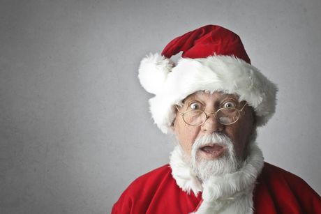 Santa Claus with an open-mouthed expression, wearing a red and white suit and hat, round glasses, and a long white beard against a grey background.