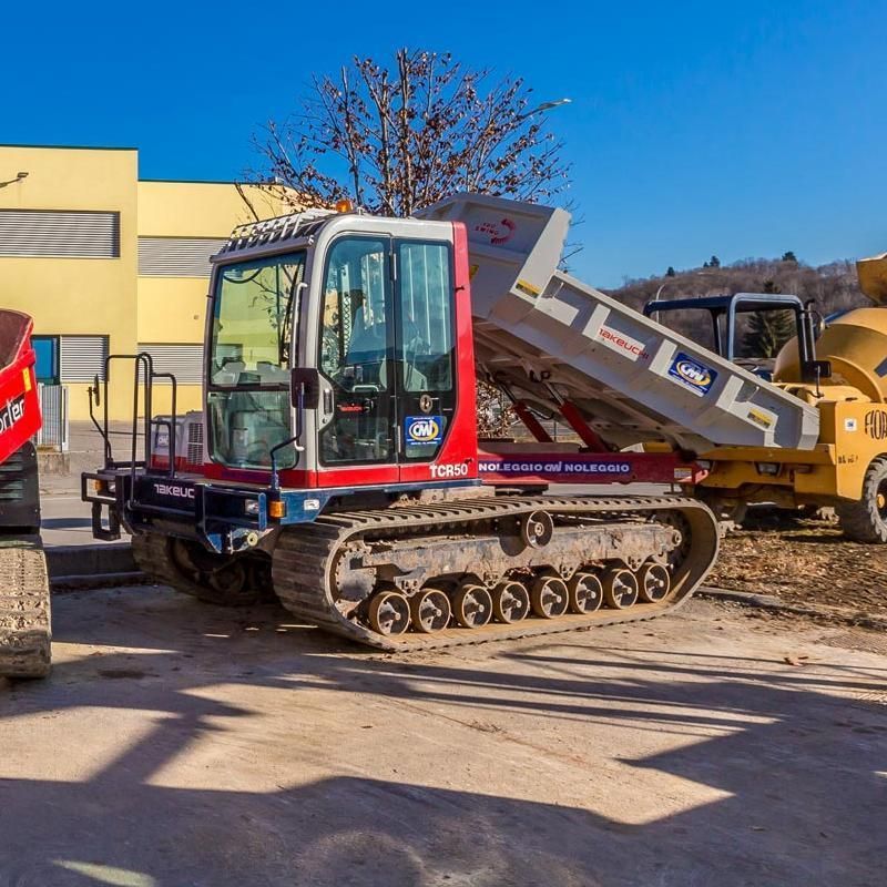 Dumper cingolato rosso e bianco con cassone rialzato, parcheggiato su cemento. Sullo sfondo un edificio giallo e altri veicoli da cantiere.
