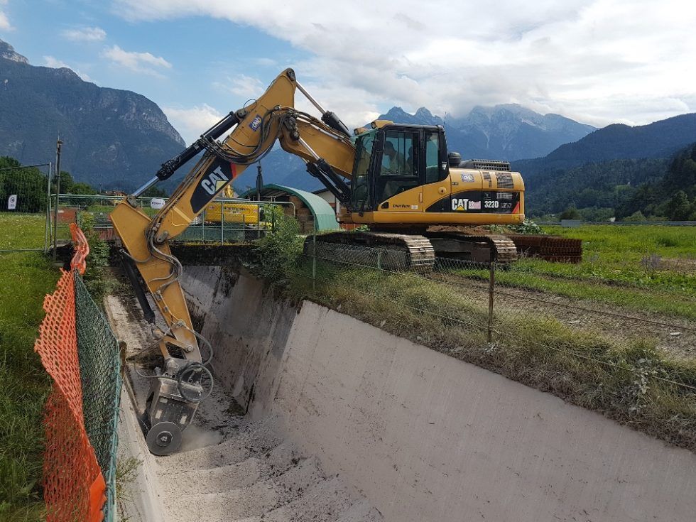 Un escavatore Caterpillar giallo scava un fossato di cemento con montagne sullo sfondo.