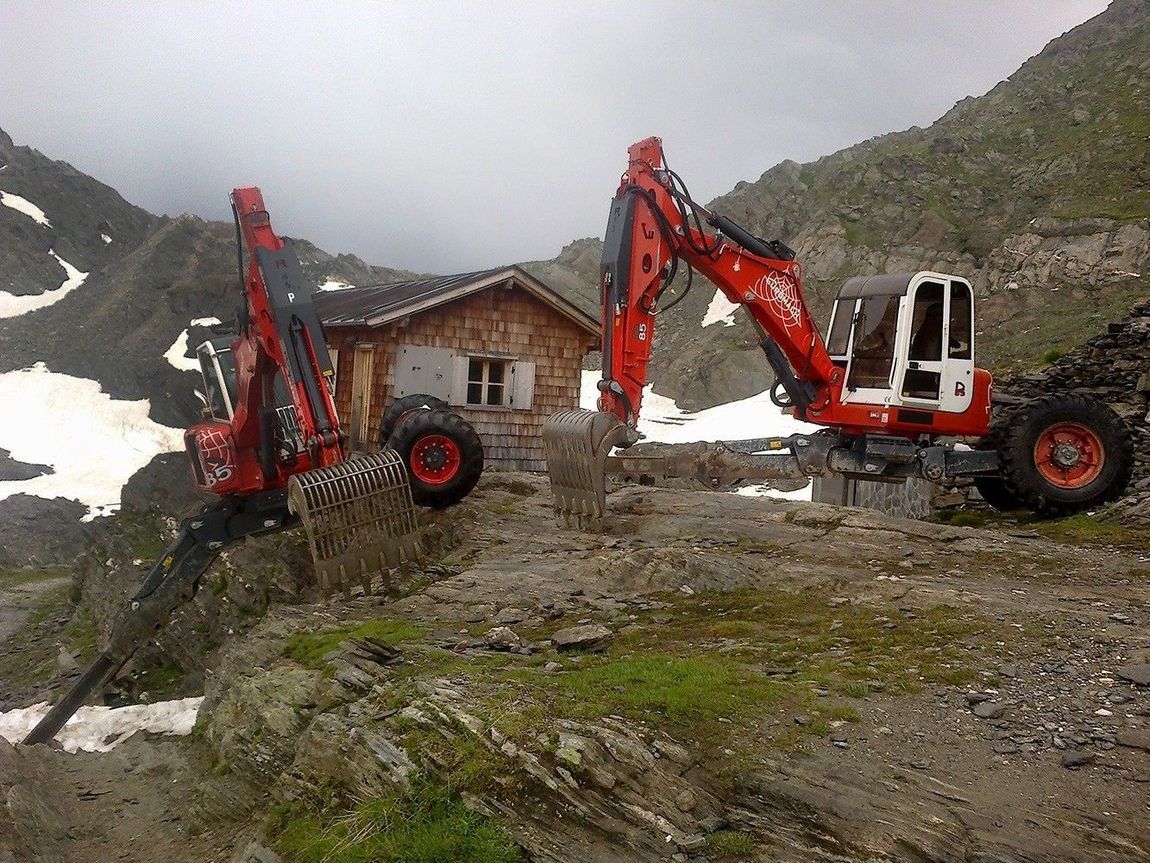 Due veicoli edili rossi lavorano vicino a un piccolo edificio in legno in un ambiente montuoso e innevato.