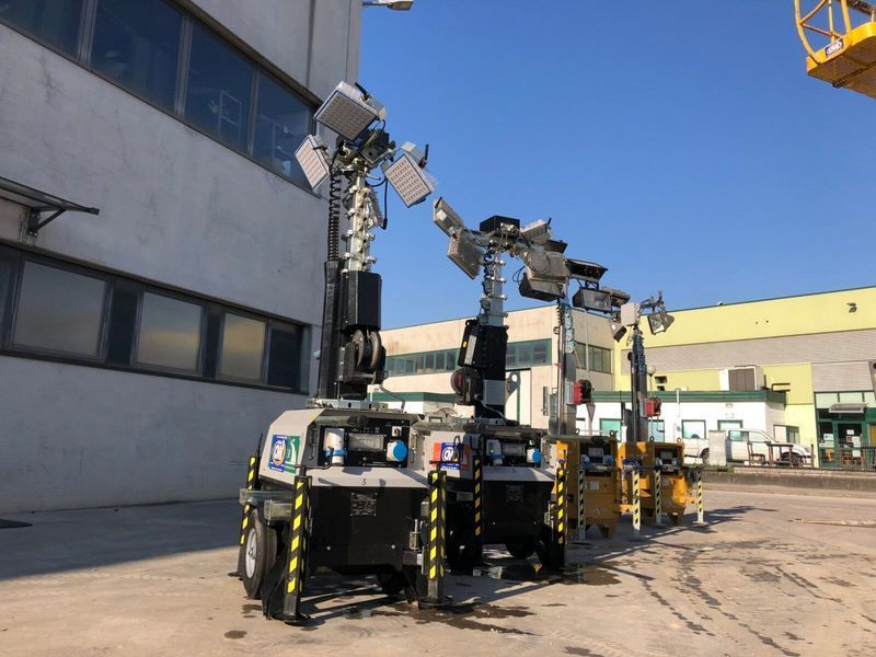 Torri faro mobili parcheggiate all'esterno di un edificio. Sono visibili diverse torri con gruppi ottici estesi, con segnaletica di sicurezza gialla e nera.