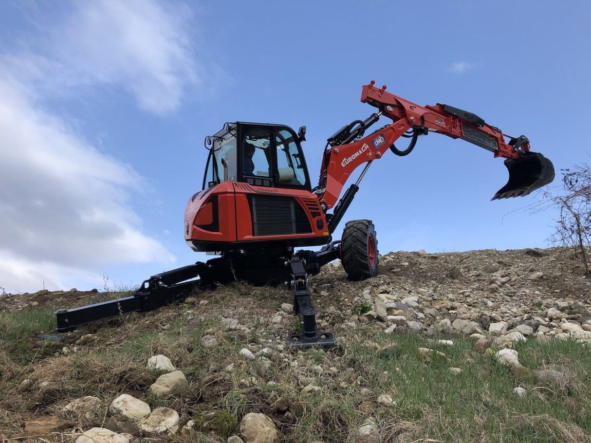 Un escavatore rosso su una collina con un cielo azzurro. Il braccio è esteso, pronto a scavare.