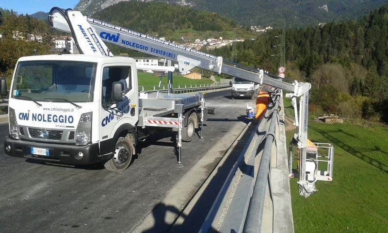 Camion con braccio sollevato che lavora sulla ringhiera di un ponte; montagne sullo sfondo.