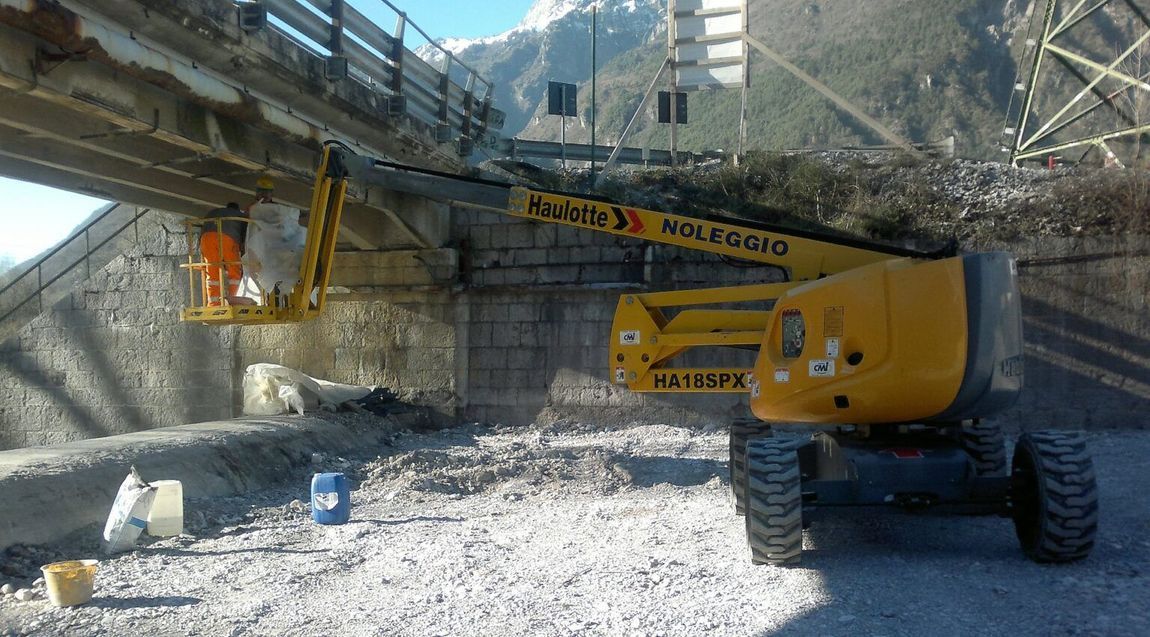 Gru gialla sotto un ponte. Due operai con gilet arancioni al lavoro. Sullo sfondo montagne.