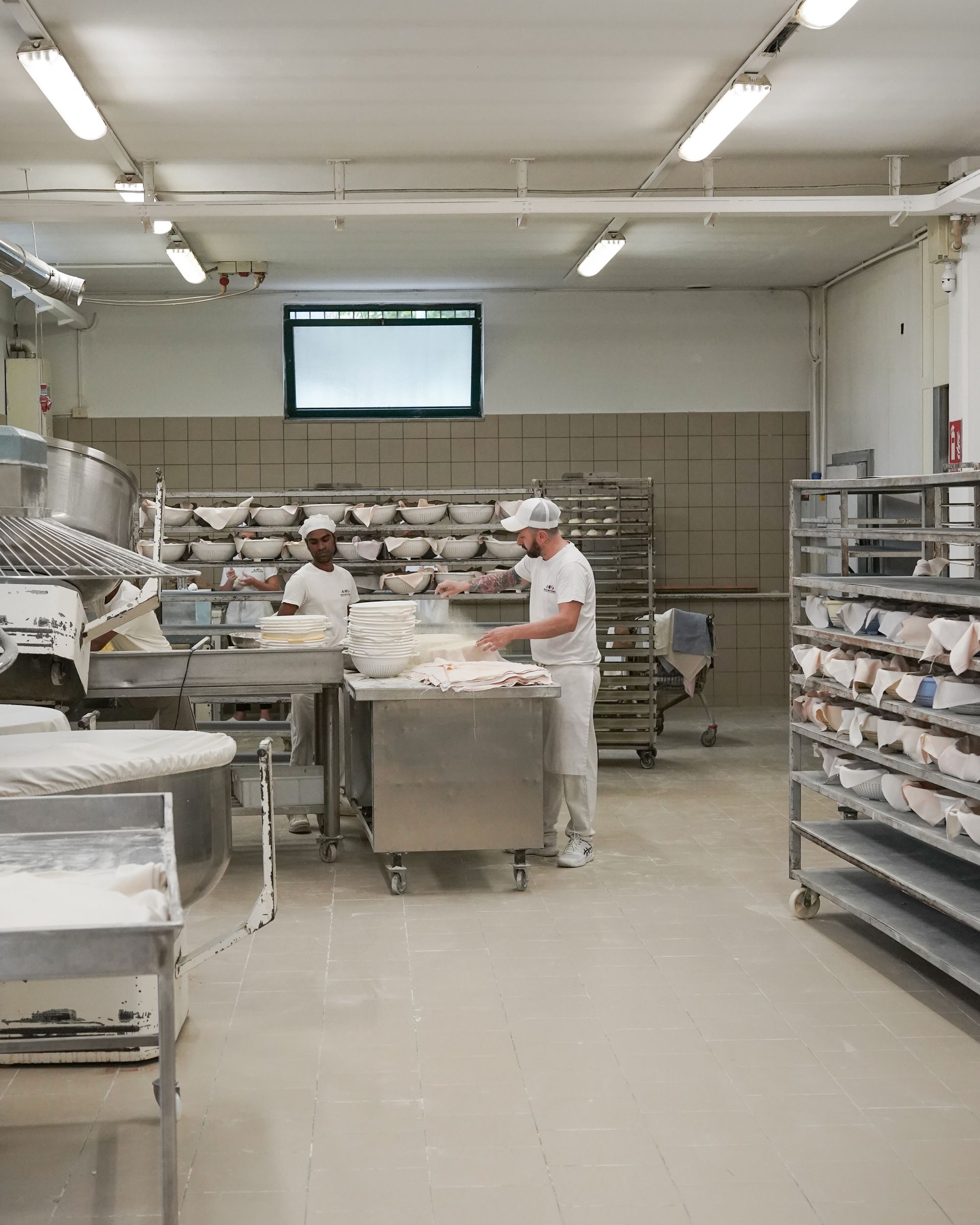 Due fornai lavorano in una cucina commerciale con attrezzature e ripiani in acciaio inossidabile; farina bianca visibile.