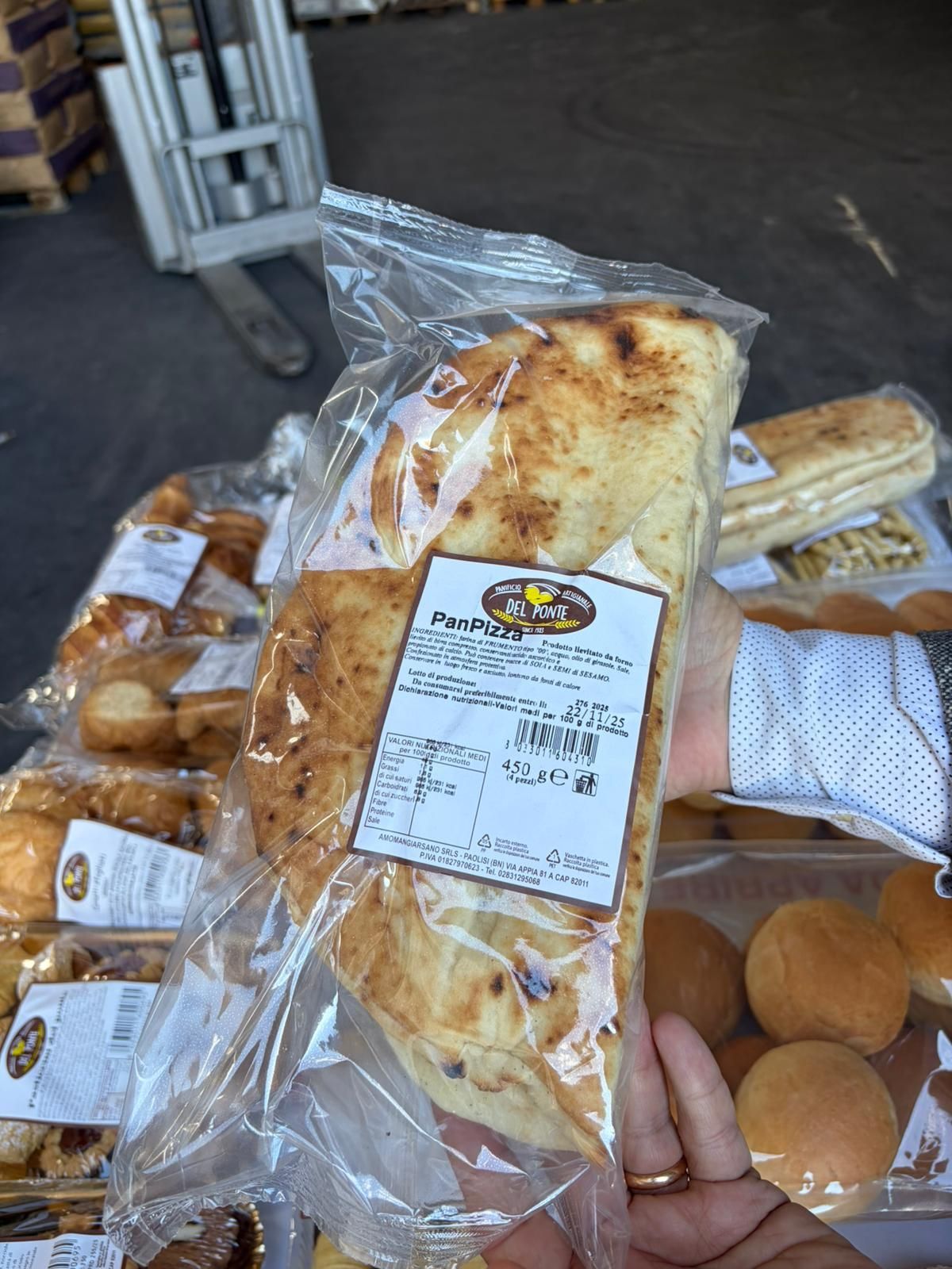 Persona che tiene in mano una confezione sigillata di pane azzimo. Altri prodotti da forno sullo sfondo, vicino a un transpallet.