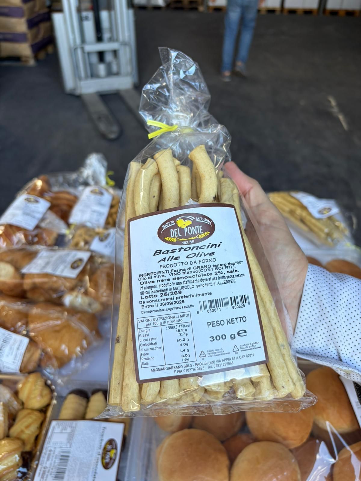 Una mano che regge un sacchetto trasparente di grissini, con l'etichetta del prodotto. Altri prodotti da forno sullo sfondo.