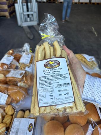 Una mano che regge un sacchetto trasparente di grissini, con l'etichetta del prodotto. Altri prodotti da forno sullo sfondo.