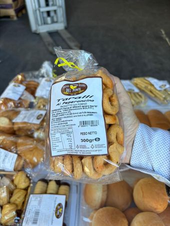 Mano che regge un sacchetto di taralli su una bancarella del mercato. Sono visibili altri prodotti da forno e un carrello elevatore.