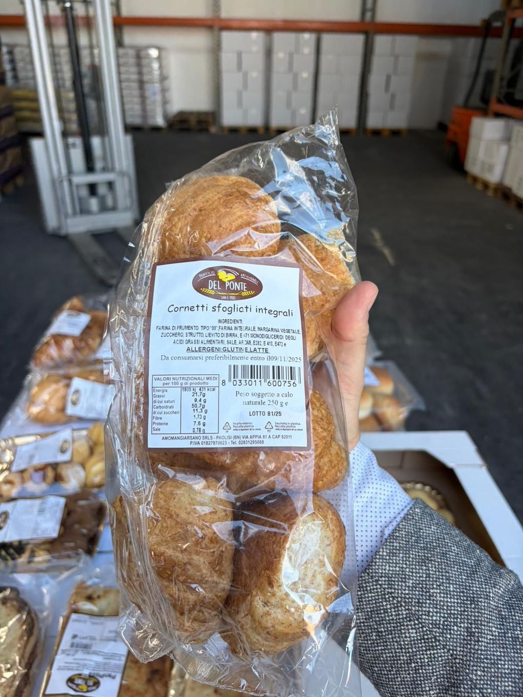 Persona che tiene in mano un sacchetto trasparente di panini con un'etichetta, probabilmente in un magazzino di una panetteria.