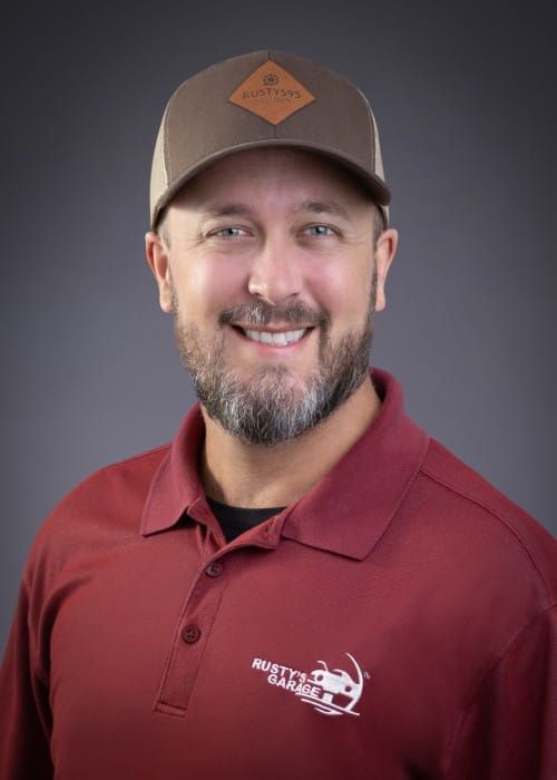 Keith smiling, wearing a red shirt with a Rusty's Garage logo, against a gray backdrop.