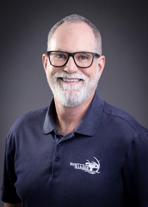 Pat smiling, wearing a blue shirt with a Rusty's Garage logo, against a gray backdrop.