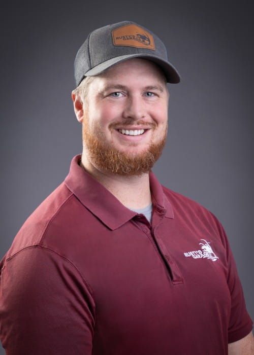 Paul smiling, wearing a red shirt with a Rusty's Garage logo, against a gray backdrop.
