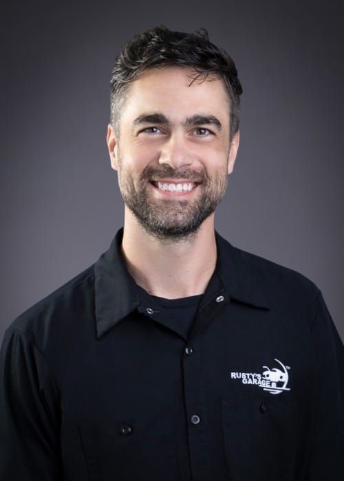 Scott smiling, wearing a black shirt with a Rusty's Garage logo, against a gray backdrop.