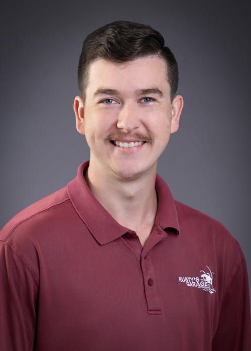 Tanner smiling, wearing a red shirt with a Rusty's Garage logo, against a gray backdrop.