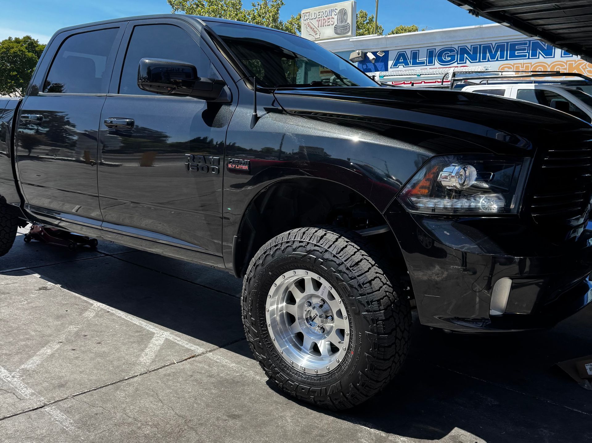 A lifted black RAM 1500 truck with a 5.7L HEMI engine, featuring polished off-road wheels and aggressive all-terrain tires, parked in front of Calderon’s Tires with signage for alignment, shocks, and other services visible in the background.
