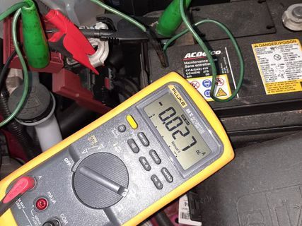 Multimeter testing car battery for parasitic draw, displaying -0.027 amps. Green and red cables connected. | Lains Auto Service