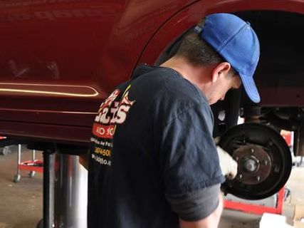 Mechanic working on car brakes; red car elevated on lift. Mechanic wears blue hat and black shirt. | Lains Auto Service