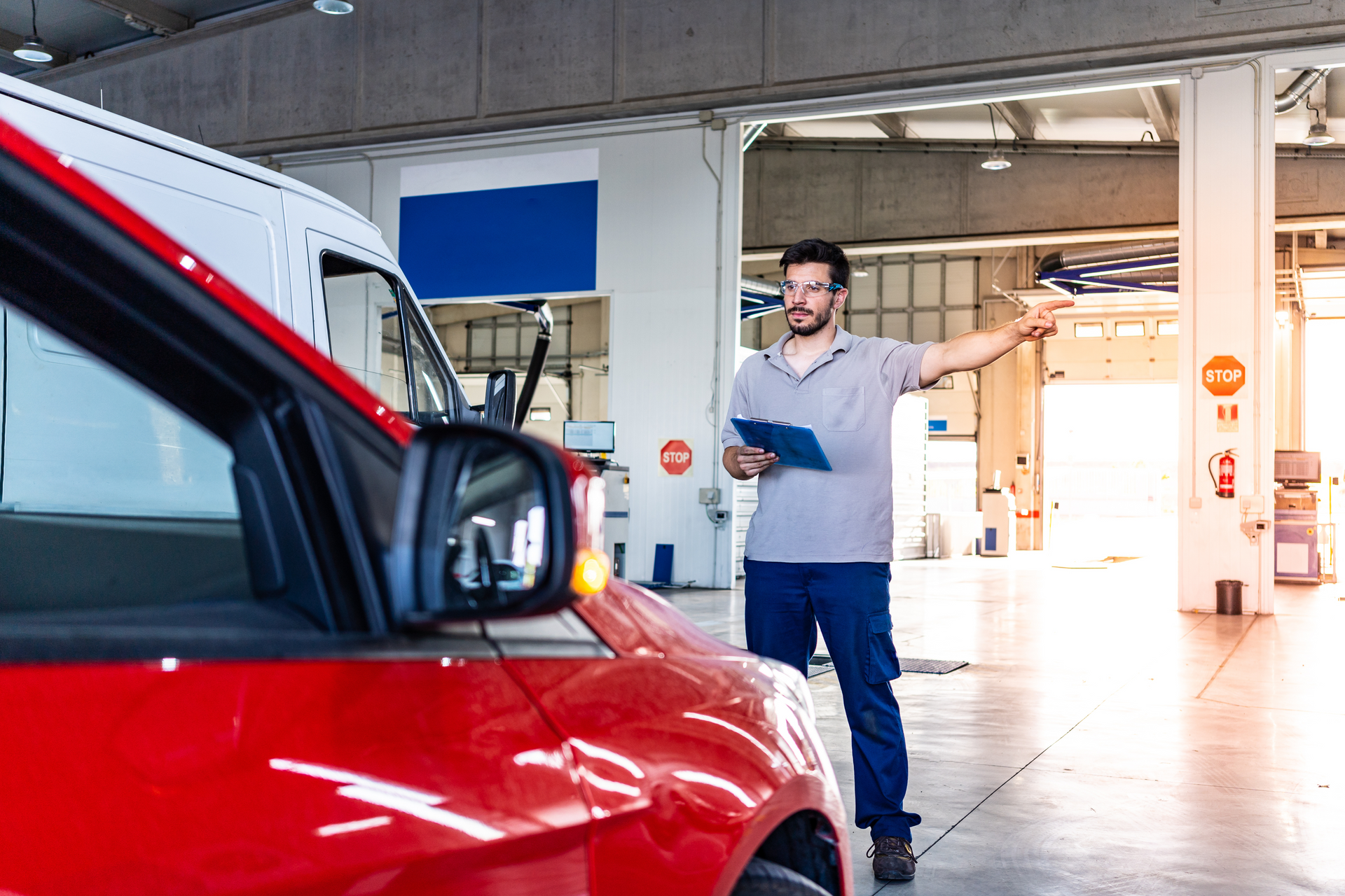 Mechanic pointing, holding clipboard, standing near red car in auto shop. | Lains Auto Services
